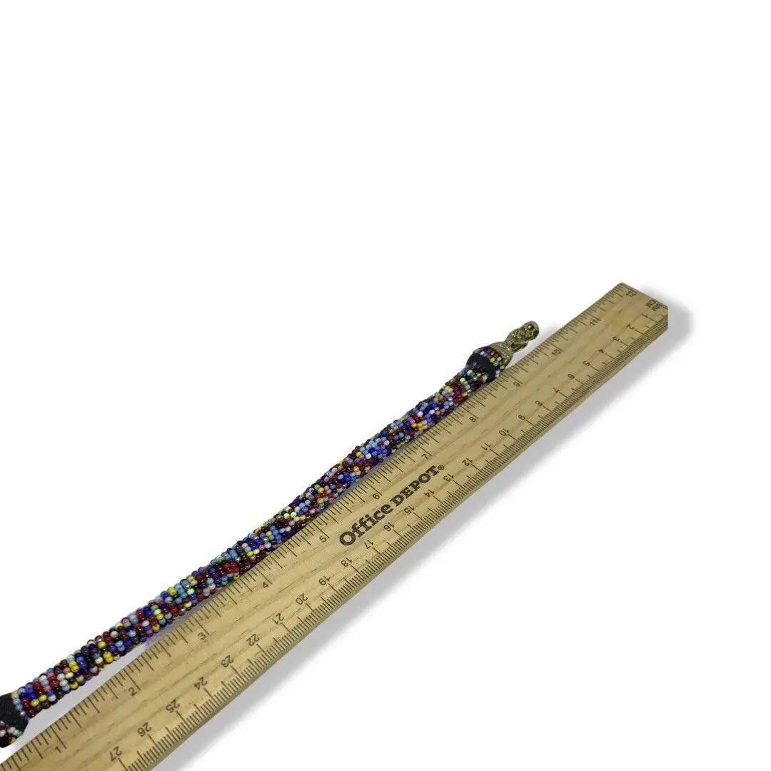 The SanteriaGuide "Ilde/Bracelet/Manilla de Diferentes Orishas/Santos con Bronze: Elegancia y Resistencia," rich in multicolored beads and reflecting the vibrant styles of various Orishas, is placed next to a wooden Office Depot ruler. The ruler showcases the bracelet’s length as it extends slightly past the 6-inch mark against a plain white background.