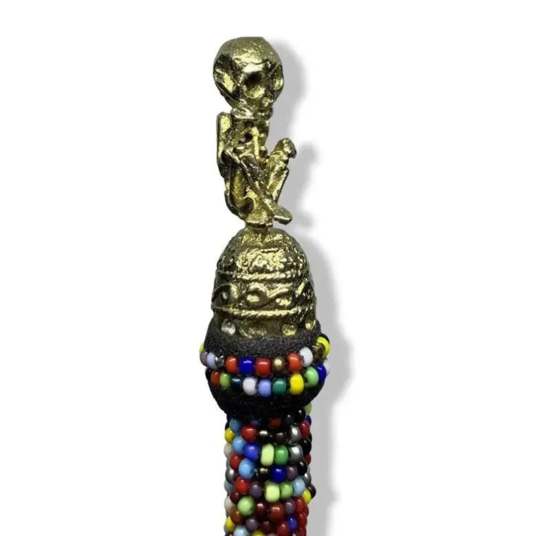 A decorative staff features a golden skeletal figure perched on top. Below the figure, a rounded section adorned with colorful beads in various hues—red, blue, yellow, and green—like those found in the SanteriaGuide Ilde / Bracelet / Manilla de Diferentes Orishas/Santos con Bronze: Elegancia y Resistencia encircles the staff. The background is plain white.